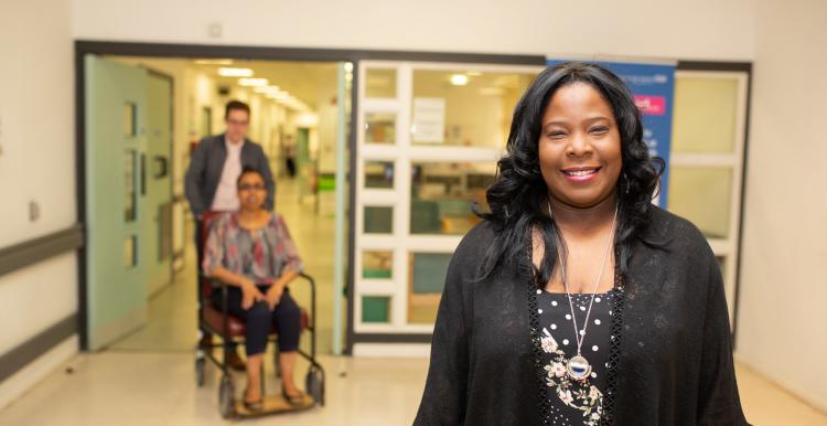 Woman in hospital smiling at the camera with a man pushing a woman in a wheelchair in the background